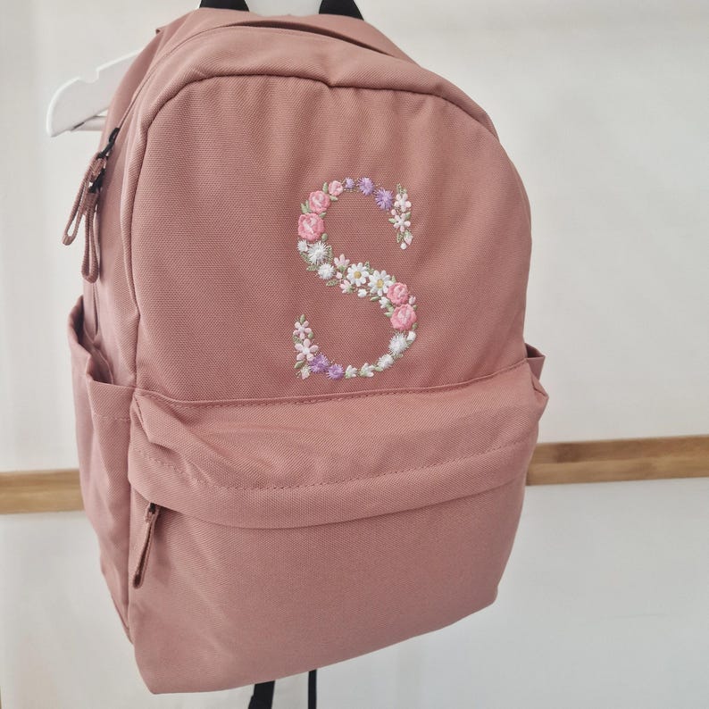 Puede incluir: Una mochila rosa polvoriento con una "S" floral bordada en la parte delantera. La mochila tiene un bolsillo delantero, un bolsillo lateral y una cremallera superior. La letra bordada est&aacute; hecha de flores rosas, blancas y moradas.