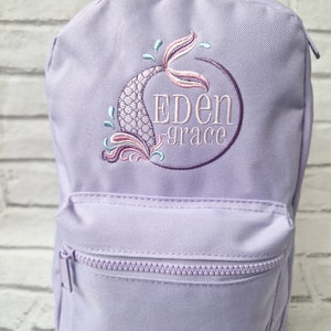 May include: A lavender backpack with a front zippered pocket. The backpack features an embroidered mermaid tail design in shades of pink, purple, and teal, with the name "Eden Grace" embroidered in purple within a circle.