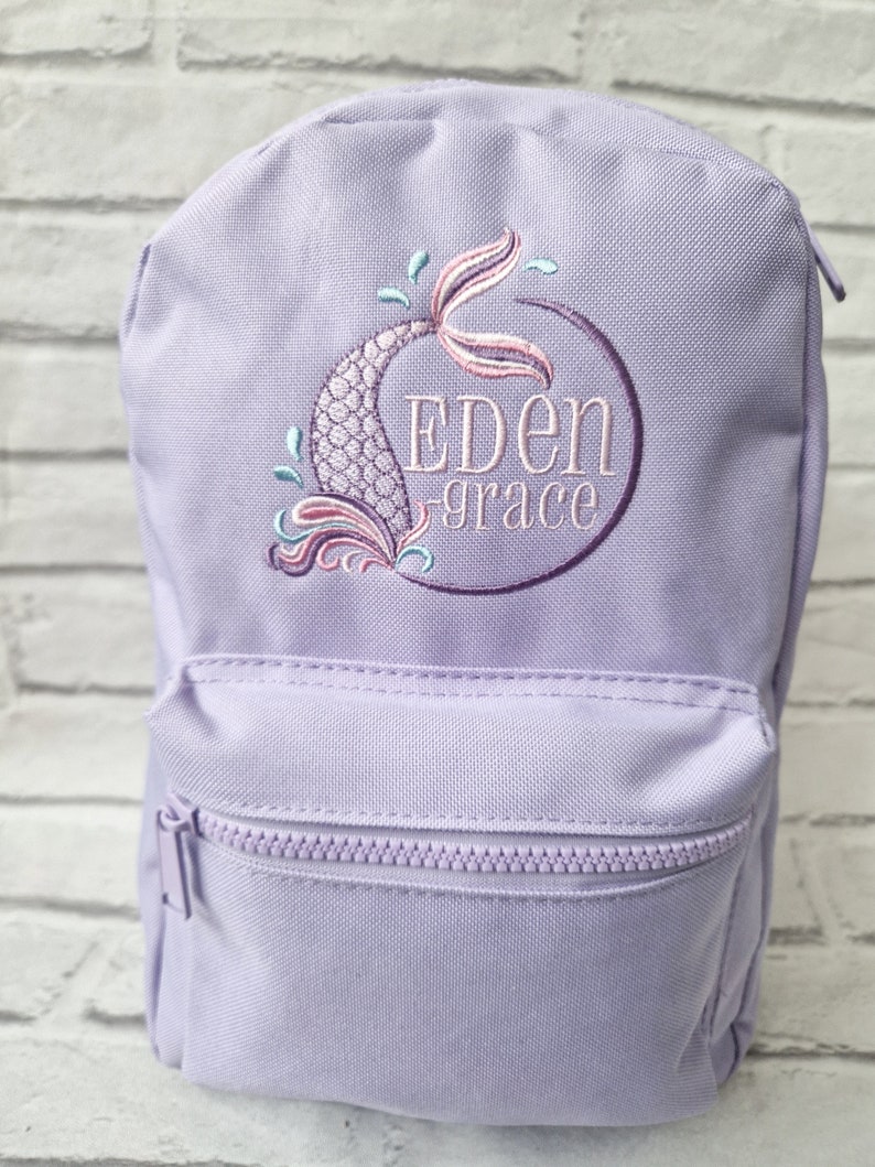 May include: A lavender backpack with a front zippered pocket. The backpack features an embroidered mermaid tail design in shades of pink, purple, and teal, with the name "Eden Grace" embroidered in purple within a circle.