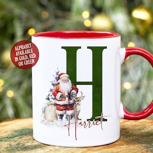 May include: White ceramic mug with a red rim and handle. The mug features a green letter "H" with a Santa Claus and reindeer illustration. The name "Harriet" is written below the letter.