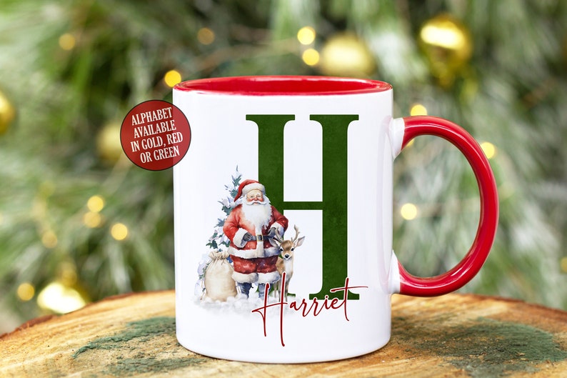 May include: White ceramic mug with a red rim and handle. The mug features a green letter "H" with a Santa Claus and reindeer illustration. The name "Harriet" is written below the letter.