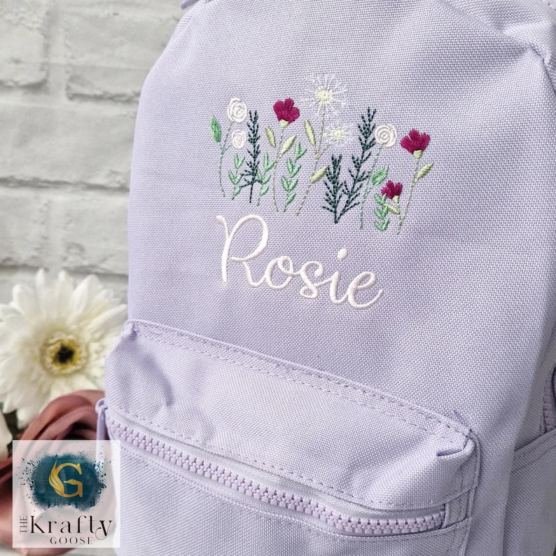 May include: A lavender backpack with a front zippered pocket. The backpack features an embroidered floral design in white, green, and magenta. The name "Rosie" is embroidered in white script below the flowers.
