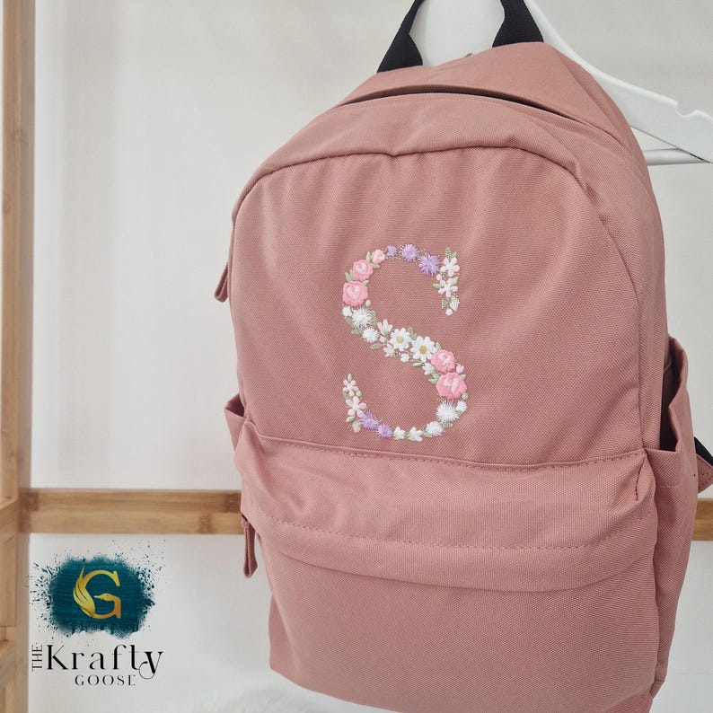 Puede incluir: Mochila rosa empolvado con una 'S' bordada floral en la parte delantera. El bordado floral incluye flores rosas, blancas y moradas. La mochila tiene un bolsillo frontal, bolsillos laterales y correas negras.
