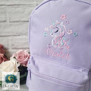 May include: A lilac backpack with a front zipped pocket. It features an embroidered unicorn design in pink, blue, and white, and the name "Violet" in pink script. The unicorn is surrounded by embroidered stars in various pastel colours.