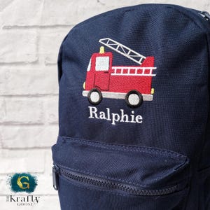 May include: A navy blue backpack featuring an embroidered red fire engine design. The fire engine has a white ladder and the name "Ralphie" embroidered in white. The bag has a front zip pocket.