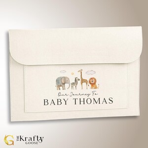 May include: A white felt envelope with a pocket on the front. The pocket has a design of a line of safari animals including an elephant, zebra, giraffe, and lion. The text "Our Journey To BABY THOMAS" is printed below the animals.