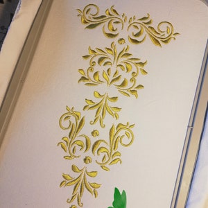 May include: Close-up of an embroidery project featuring a gold floral design on a white fabric. The design includes swirling patterns and leaf-like shapes. The text "Amalithya The Art of Costumes" is visible.