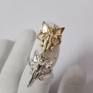 May include: Two rings, one gold and one silver, each with a fairy design. The fairies have detailed wings and are gracefully posed. The rings are shown against a white background.