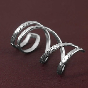 May include: A silver-coloured, spiral ring with a textured, engraved design. The ring curves in a continuous loop, with detailed floral or leaf patterns. The metal has a polished finish, reflecting light against a dark red background.