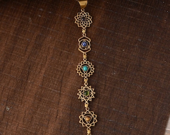 Può includere: Una collana a catena dorata con sette simboli dei chakra, ognuno con una pietra preziosa di colore diverso. I simboli sono collegati da piccole perle dorate. La collana è su una superficie di legno marrone.