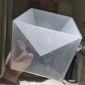 Puede incluir: Un recipiente cuadrado de plástico transparente con una abertura triangular en la parte superior. El recipiente está hecho de un material translúcido y tiene una superficie lisa.