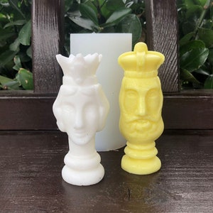 May include: Two white and yellow ceramic chess pieces, a king and a queen, with detailed faces and crowns. The pieces are on a brown wooden surface.