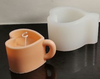 Può includere: Una candela a forma di cuore color pesca con uno stoppino bianco e un manico. La candela è accanto a uno stampo in silicone bianco a forma di cuore con un manico.