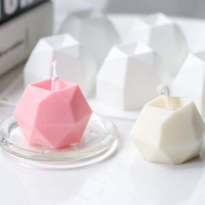 Puede incluir: Una vela geométrica rosa sobre un plato de vidrio transparente, junto a una vela geométrica blanca. Varios moldes de velas geométricas blancas están en el fondo. Las velas tienen mechas y están sobre una superficie blanca.