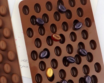 May include: Two brown silicone molds for making chocolate. The mold on the left has 42 individual cavities shaped like coffee beans. The mold on the right has 42 individual cavities shaped like coffee beans, and is filled with various colored chocolate candies.