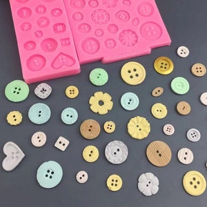 Op de afbeelding: Een roze siliconen mal met twee delen, elk met verschillende knoopvormen. De mal ligt naast een verzameling kleurrijke, decoratieve knopen.