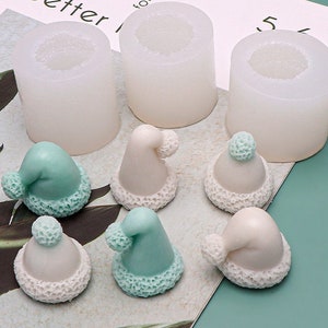 Könnte beinhalten: Drei weiße Silikonformen zum Herstellen kleiner Weihnachtsmützen. Sechs fertige Mützen sind abgebildet, drei in Weiß mit einem hellblauen Bommel und drei in Hellblau mit einem weißen Bommel.
