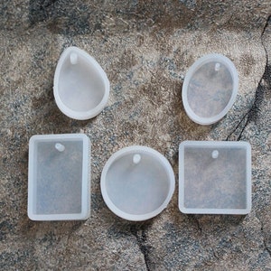 Op de afbeelding: Vijf transparante siliconen mallen voor het maken van sieraden hangers. De mallen zijn in verschillende vormen, waaronder een cirkel, ovaal, rechthoek en traan. Elke mal heeft een klein gaatje bovenaan om een ​​beugel aan te bevestigen.