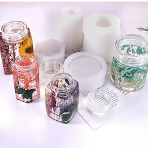 Puede incluir: Seis frascos de vidrio transparente con diferentes flores y hierbas secas en su interior. Los frascos están sobre una superficie blanca con moldes de silicona blanca para hacer manualidades con resina.