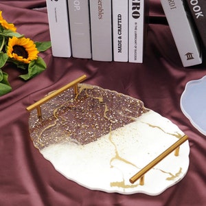 Könnte beinhalten: Ein weißes und goldenes Serviertablett aus Harz mit einer einzigartigen, organischen Form und zwei goldenen Metallgriffen. Das Tablett hat ein marmoriertes Design mit goldenen Akzenten und einem glitzernden Finish.
