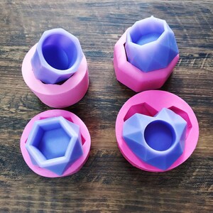 May include: Four silicone molds in pink and purple. The molds are in the shape of geometric designs. Two are round, one is heart-shaped, and one is a geometric shape. The molds are on a wooden surface.