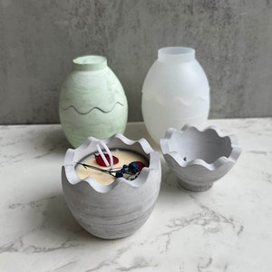 May include: Three concrete egg-shaped candle holders. Two are empty and one is filled with a white candle with red and blue accents. The candle holders are on a white marble surface.