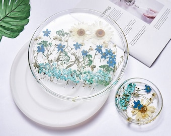 May include: Two clear resin trays with dried flowers and leaves embedded inside. The larger tray is on a white surface and the smaller tray is on top of the larger tray. The flowers are white, blue, and green.