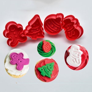 Op de afbeelding: Set van vier rode plastic kerstkoekjesvormen in de vorm van een gingerbread man, een kerstboom, een ornament en een sneeuwpop. De vormen worden getoond naast vier cupcakes die zijn versierd met glazuur en dezelfde vormen.
