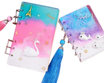 Può includere: Due piccoli quaderni a spirale, colorati, con dettagli di glitter e resina. Un quaderno presenta un cigno bianco con una corona dorata e il testo "Scorpio". L'altro quaderno presenta un delfino bianco con una corona dorata e il testo "Angel". Entrambi i quaderni hanno nappine blu.
