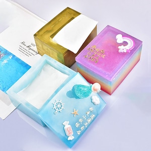 Könnte beinhalten: Drei dekorative Taschentuchboxen mit verschiedenen Designs. Die erste Box ist golden mit einem weißen Taschentuch im Inneren. Die zweite Box ist ein Regenbogen-Verlauf mit einer rosa Oberseite und einem Regenbogen-Design. Die dritte Box ist hellblau mit einem weißen Taschentuch im Inneren und einem Meerjungfrauen-Design auf dem Deckel. Der Deckel ist geöffnet und das Taschentuch ist sichtbar.