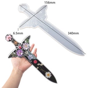 Könnte beinhalten: Eine transparente Silikonform in Form eines Schwertes mit einem Schmetterlingsdesign. Die Form ist 39,6 cm lang und 16,5 cm breit. Die Form wird neben einem fertigen Schwert gezeigt, das mit gepressten Blumen hergestellt wurde.