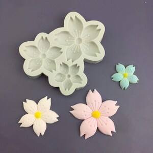 May include: Silicone mold for making cherry blossom shaped fondant decorations. The mold has three different sized blossoms. There are also three finished fondant blossoms in pink, white, and blue.
