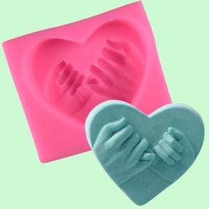 May include: Pink silicone mould shaped like a heart with two hands holding pinkies together. A teal blue heart-shaped cake with two hands holding pinkies together is next to the mould.