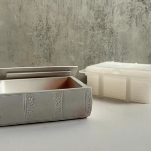 May include: A white ceramic book-shaped box with a lid open, revealing an empty interior. The box has decorative details on the front and sides. A second white ceramic box with a lid is also shown, but it is closed.