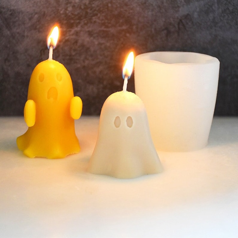 May include: Two ghost-shaped candles, one yellow and one white, with wicks and flames. A white silicone mold for making ghost candles is also shown.