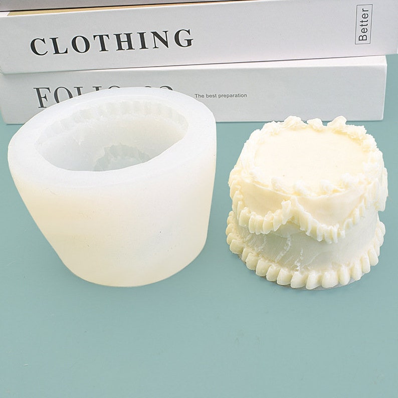 Peut inclure: Un moule en silicone blanc pour le bricolage, &agrave; c&ocirc;t&eacute; d'un g&acirc;teau &agrave; plusieurs &eacute;tages, blanc cass&eacute;. Le moule est cylindrique avec un bord festonn&eacute;. Le g&acirc;teau a un design similaire, avec plusieurs couches et un dessus d&eacute;coratif.