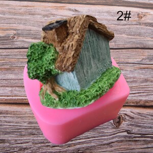 Puede incluir: Molde de silicona rosa en forma de coraz&oacute;n, con un dise&ntilde;o 3D detallado de una peque&ntilde;a casa con un &aacute;rbol que crece en el techo. La casa est&aacute; pintada de azul y verde, con ramas de &aacute;rbol marrones. El molde est&aacute; etiquetado con "2#" en la esquina superior derecha.