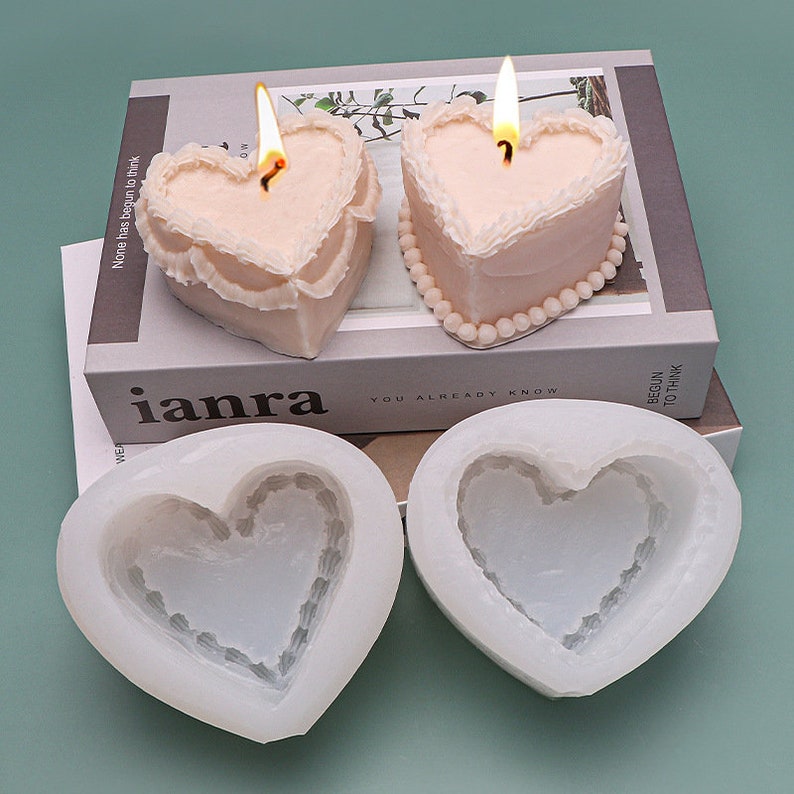 K&ouml;nnte beinhalten: Zwei rosa herzf&ouml;rmige Kerzen mit wei&szlig;em Frosting-Design und wei&szlig;em Perlenrand. Zwei herzf&ouml;rmige Silikonformen mit wei&szlig;em Frosting-Design.