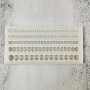 May include: A white silicone mold with multiple rows of various sized circular and scalloped designs. The mold is rectangular and sits on a light gray surface. The designs are for creating decorative elements.