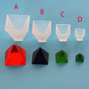 Può includere: Quattro stampi in silicone trasparente di diverse dimensioni, etichettati A, B, C e D. Ogni stampo ha la forma di una piramide quadrata. Sotto gli stampi sono mostrate quattro piramidi in resina finite: rossa, nera, verde e una verde più piccola.