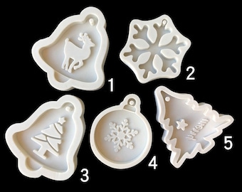 Puede incluir: Cinco moldes de silicona blancos para hacer adornos navideños. Los moldes tienen forma de campana con un reno, un copo de nieve, una campana con un árbol de Navidad, un adorno redondo con un copo de nieve y un árbol de Navidad con una estrella.