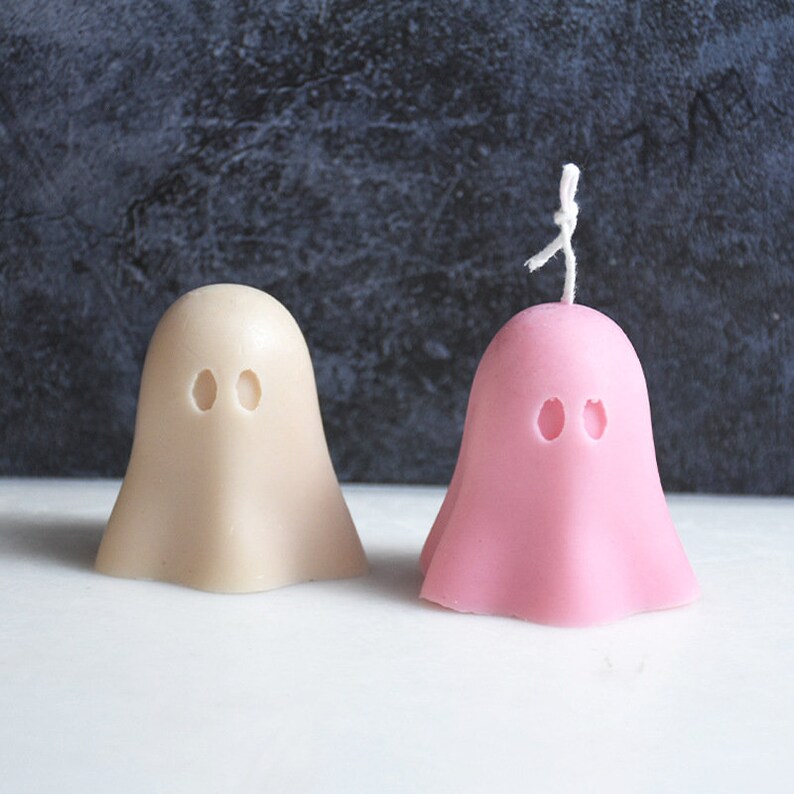 May include: Two ghost-shaped candles, one in a light beige color and the other in a light pink color. Both candles have two black eye holes and a white wick.