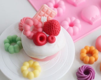 May include: A white cake with pink frosting decorated with a chocolate bar, strawberries, and red and pink donut-shaped candies. The cake is on a white cake stand with a green donut, a yellow donut, and a purple donut on the table.