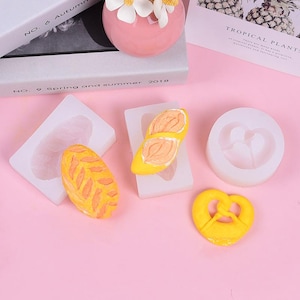 May include: Four silicone moulds for baking, shaped like a pretzel, a leaf, a butterfly, and a heart. The moulds are white and the baked goods are yellow.