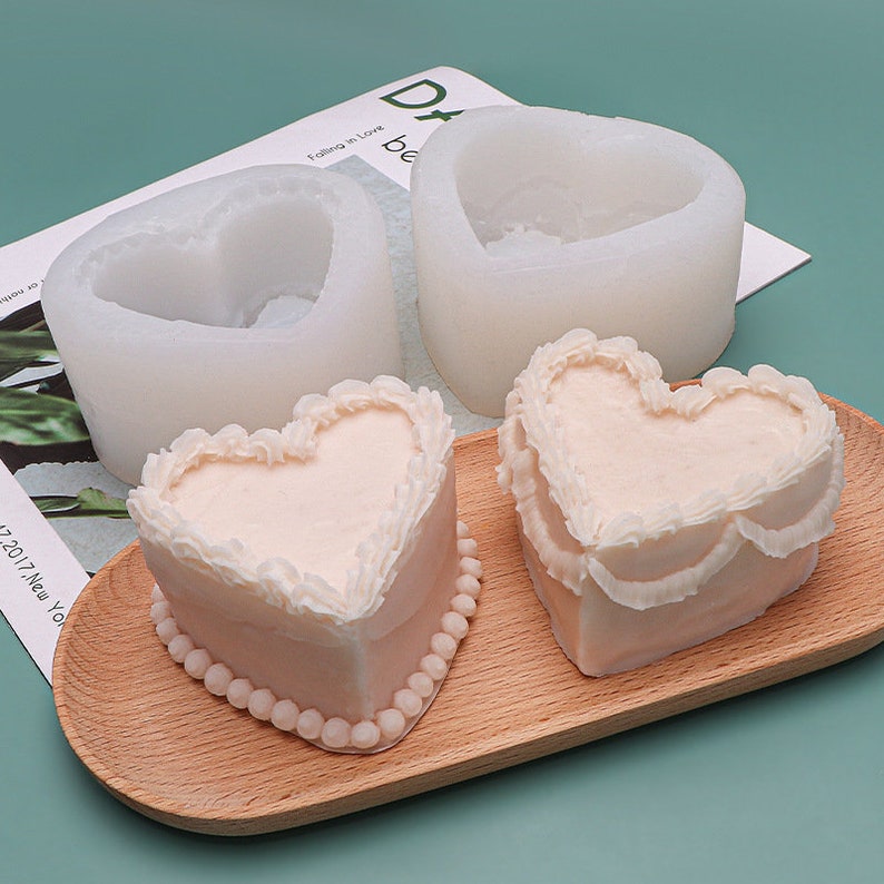 K&ouml;nnte beinhalten: Zwei wei&szlig;e Silikonformen in Herzform und zwei herzf&ouml;rmige Kuchen, die mit wei&szlig;em Frosting und perlenartigen Akzenten auf einem Holztablett verziert sind.