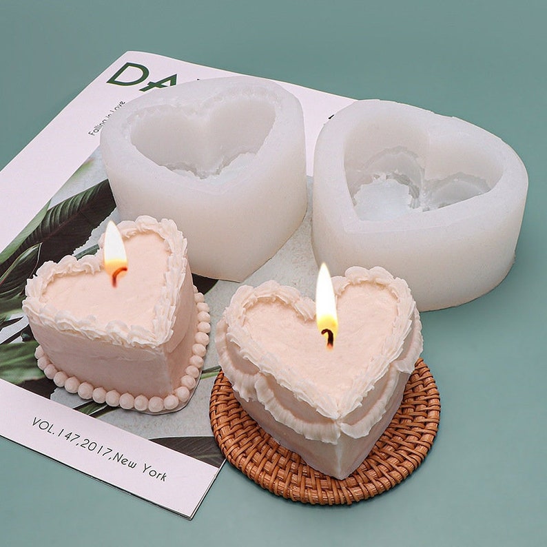 K&ouml;nnte beinhalten: Zwei herzf&ouml;rmige Silikonformen zum Herstellen von Kerzen, mit zwei fertigen herzf&ouml;rmigen Kerzen mit wei&szlig;em Frosting und einem brennenden Docht.