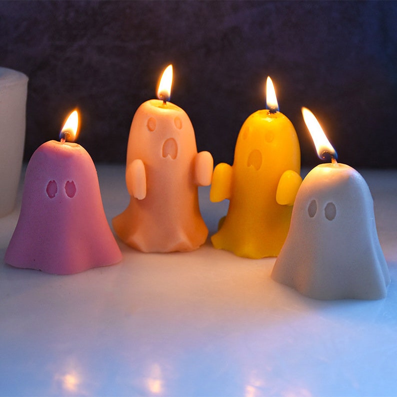 May include: Four ghost-shaped candles, one pink, one orange, one yellow, and one gray, with flames lit.