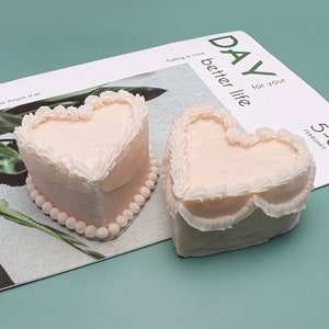 K&ouml;nnte beinhalten: Zwei herzf&ouml;rmige Kuchen, die mit wei&szlig;em Frosting und perlenartigen Akzenten verziert sind. Die Kuchen befinden sich auf einer wei&szlig;en Oberfl&auml;che mit dem aufgedruckten Text "DAY for your better life" und "Falling in Love".