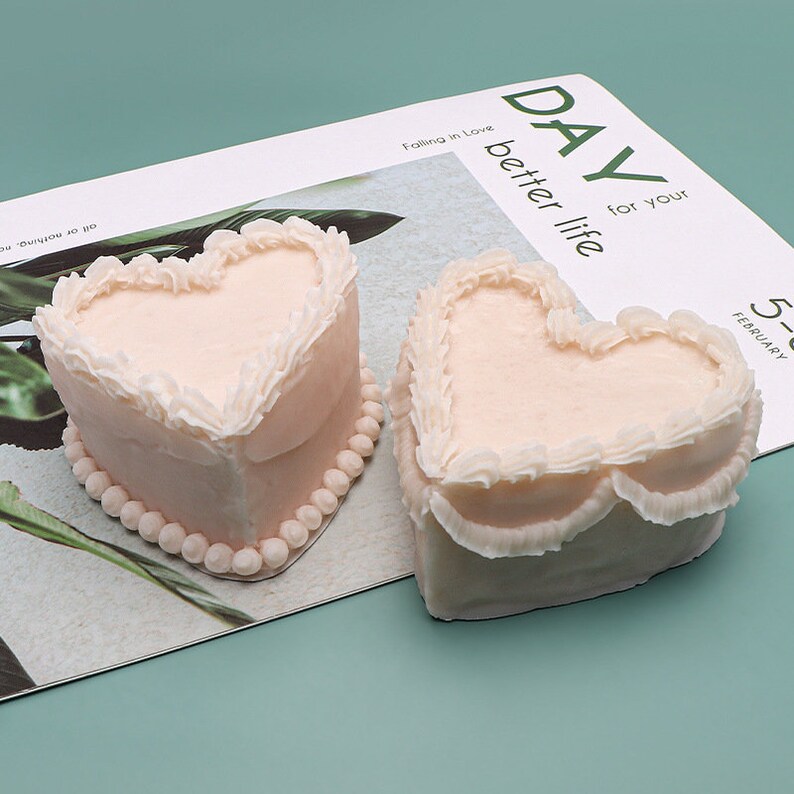 K&ouml;nnte beinhalten: Zwei herzf&ouml;rmige Kuchen, die mit wei&szlig;em Frosting und perlenartigen Akzenten verziert sind. Die Kuchen befinden sich auf einer wei&szlig;en Oberfl&auml;che mit dem aufgedruckten Text "DAY for your better life" und "Falling in Love".