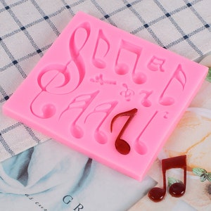 Puede incluir: Molde de silicona rosa con múltiples formas de notas musicales. El molde está sobre una superficie con estampado rosa y blanco. Hay dos notas musicales rojas en la superficie junto al molde.